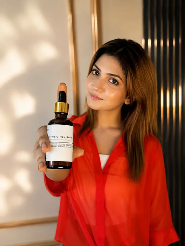 লাল শার্ট পরা তরুণী হাসিমুখে The Formulate রোজমেরি হেয়ার সিরাম হাতে ধরে আছেন